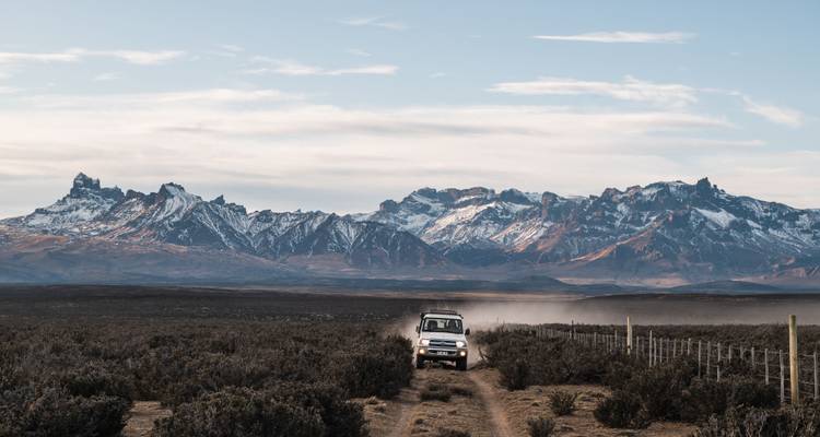 Piste poussiéreuse avec 4x4 roulant vers une vaste chaîne de montagnes déchiquetées sous un ciel pâle.