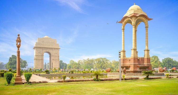 Sunny view of India Gate war memorial and neighbouring canopy monument set in manicured gardens under blue sky.