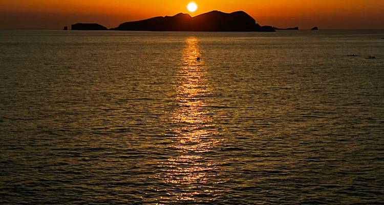 Brilliant orange sun setting behind a distant island, casting a glowing path across calm sea water.
