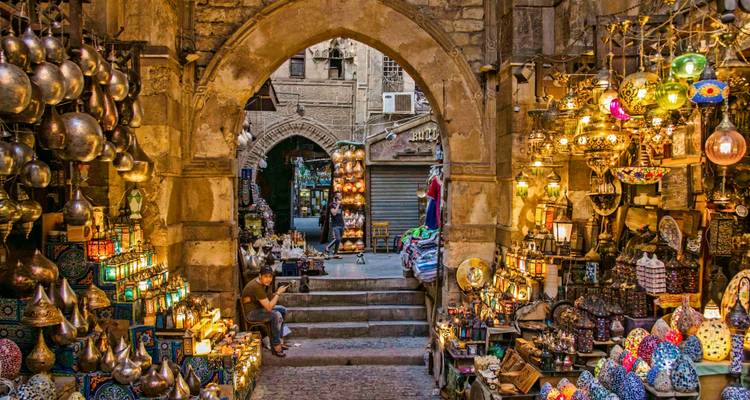 Atmospheric archway leading to a bustling souk filled with brassware, lamps and a seated vendor in Cairo.