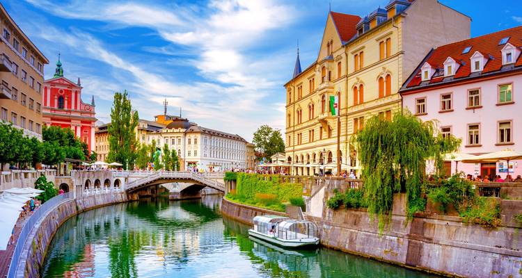 Charmant grachttafereel in Ljubljana met sierlijke brug, pastelkleurige gebouwen en een rondvaartboot onder een blauwe hemel.