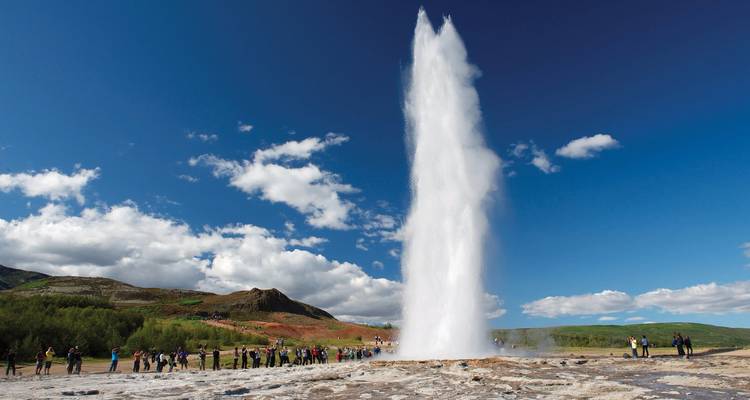 Tourists watch a geyser erupt high into the air under a bright Icelandic sky.