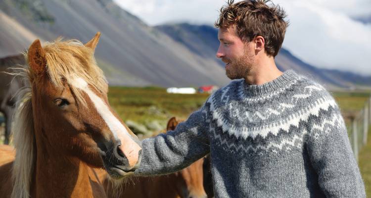 Young man in wool sweater gently strokes an Icelandic horse with rugged mountains behind.