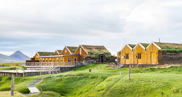 Traditional turf-roof wooden farm buildings sit on rolling green pastures beneath soft clouds.