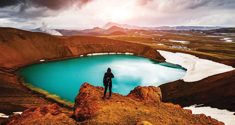 Hiker surveys a vivid turquoise crater lake from a rugged red volcanic ridge in Iceland.