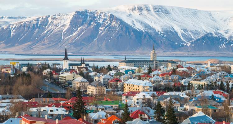 Panoramic cityscape of Reykjavik with colorful houses, church spires and a snow-capped mountain backdrop across the bay.