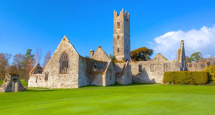 Ruines d'abbaye médiévale en pierre avec haute tour située sur un parcours de golf vert bien entretenu.