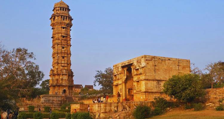 Hoge stenen Vijay Stambh toren die uitkijkt over kleine tempelstructuren en bezoekers bij Chittorgarh Fort.