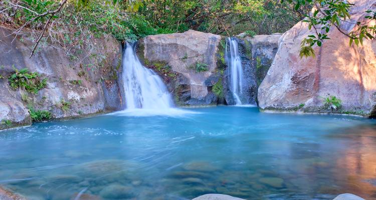 Dubbele watervallen die vallen in een serene blauwe natuurlijke poel omgeven door gladde rotswanden en weelderig gebladerte.