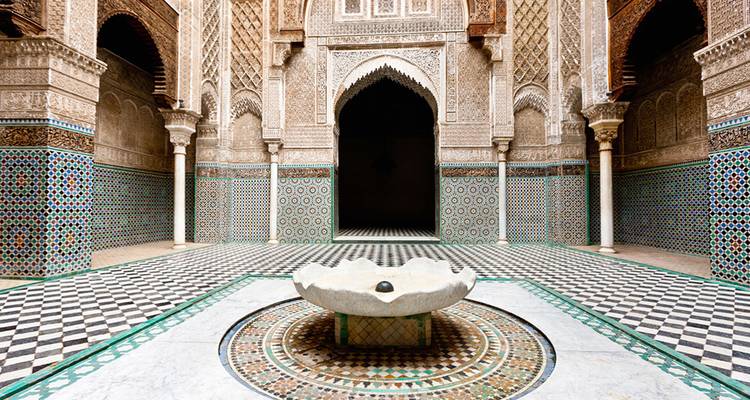 Patio intrincadamente embaldosado con fuente central rodeado de arcadas ornamentadas de una madrasa marroquí.