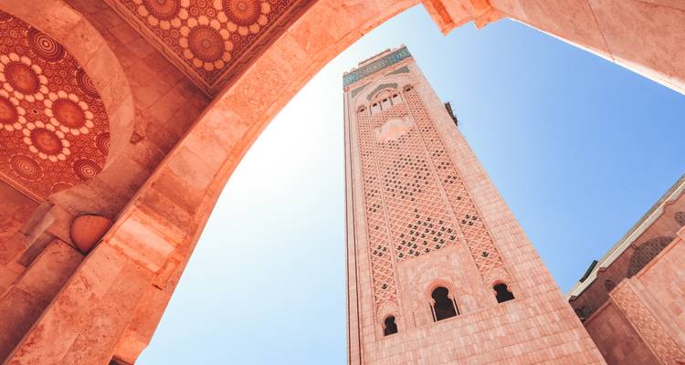 Upward view through ornate arch framing the patterned minaret of Hassan II Mosque against a bright blue sky.
