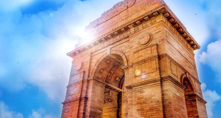 Low-angle view of India Gate in bright sunlight against a blue sky with lens flare