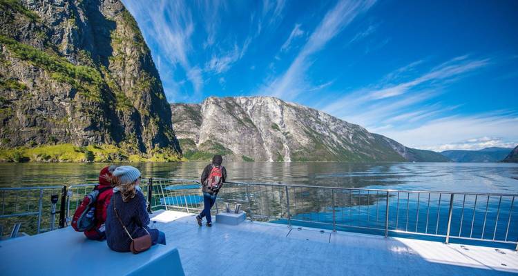Travellers enjoying the open deck of a fjord cruise ship surrounded by dramatic green cliffs and blue sky.