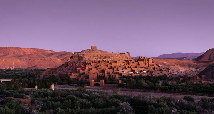 Twilight view of Aït Benhaddou bathed in purple sunset light above green oasis fields.