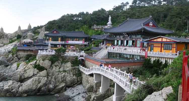 Complexe de temples bouddhistes au bord de mer perché sur des rochers escarpés avec une longue passerelle blanche et des visiteurs explorant le littoral pittoresque.