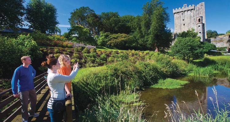 Visitors photograph landscaped gardens and tall stone tower of Blarney Castle reflected in a pond