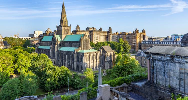 Vue en plongée à l'heure dorée de la cathédrale de Glasgow entourée d'arbres feuillus et de bâtiments urbains sous un ciel bleu dégagé.