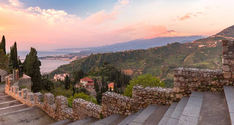 Sunrise view from Taormina terraces toward Mount Etna glowing pink above coastal valleys and Ionian Sea.