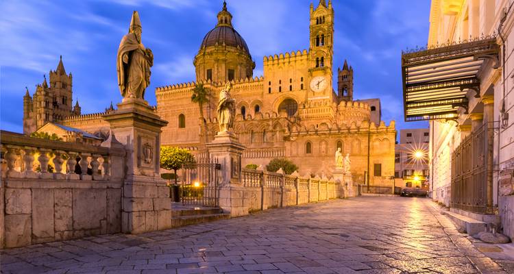 Evening lights illuminate Palermo Cathedral and statues along a cobblestone street at dusk.