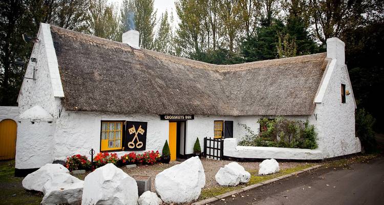 White-washed thatched Crosskeys Inn surrounded by large decorative boulders and autumn trees.