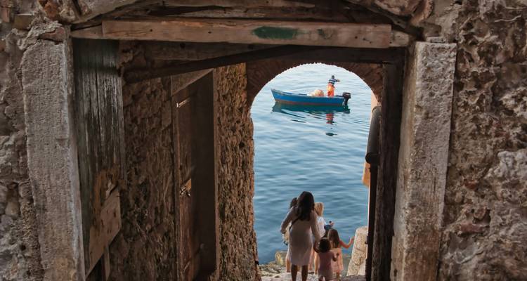 Une mère et deux enfants marchent dans un passage de pierre vers un petit bateau sur une eau bleue calme encadrée par une arche.