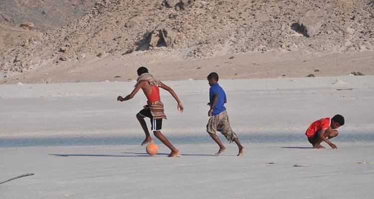 Los niños locales juegan fútbol en una playa amplia de arena blanca enmarcada por colinas rocosas.