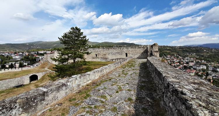 Murallas de piedra de una fortaleza en la cima de una colina que domina un pueblo moderno y colinas ondulantes.