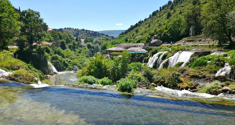 Serie de cascadas suaves que fluyen a través de un campo verdoso con ruinas de piedra cercanas.
