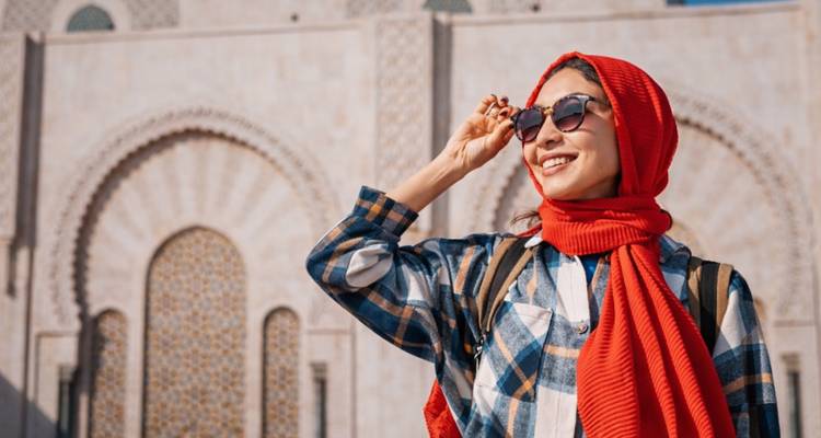 Smiling woman in sunglasses and red scarf poses before ornate stone arches of a Moroccan mosque.