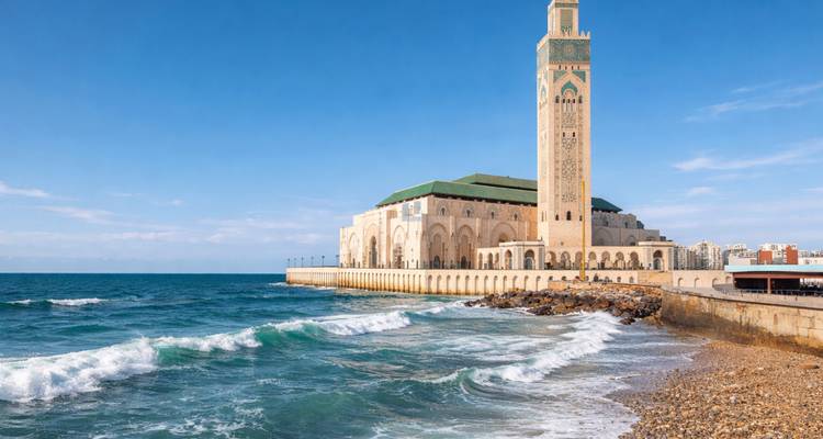 Hassan II Mosque rises beside crashing Atlantic waves under a bright blue sky.