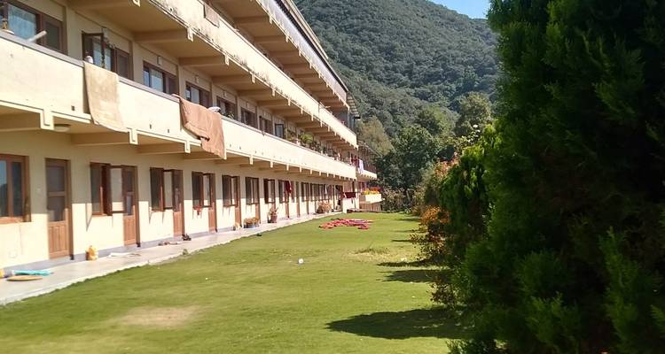Long monastic residence building with balconies facing a grassy courtyard at the foot of a forested hill.