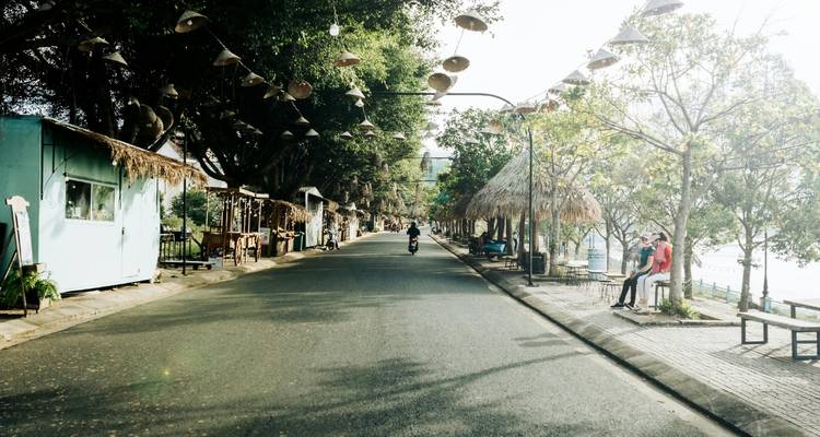 Rue calme bordée d'arbres avec de petits cafés et des jardinières suspensives décoratives, un scooter circulant au centre.