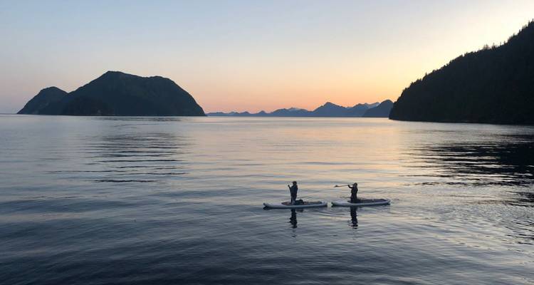 Deux pagayeurs de planche à rame glissent sur une eau miroir au crépuscule avec des îles en silhouette et un ciel pastel.