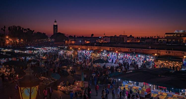 Vibrant night scene of Jemaa el-Fna square packed with bustling market stalls and glowing lights against orange sunset.