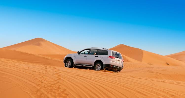 A white 4x4 SUV drives up orange sand dunes beneath a clear blue sky.