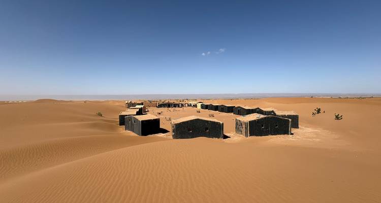Remote cluster of low black desert huts surrounded by endless sand under a vast blue sky.