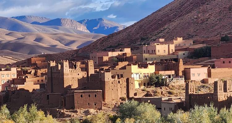 Traditional earthen village with fortified towers set against arid mountain slopes under partly cloudy skies.
