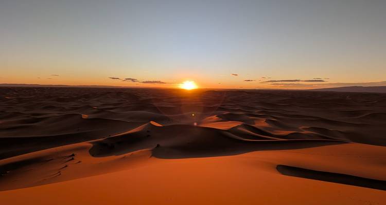 Sun sets over a vast sea of dark rolling dunes casting an orange glow on the foreground ridge.