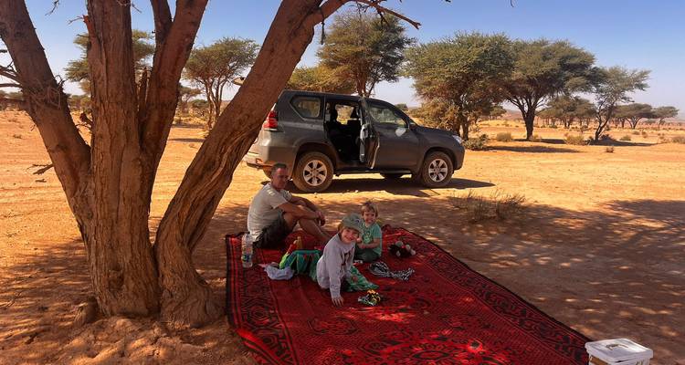Family rests on a red rug beneath an acacia tree beside their parked SUV in semi-arid landscape.