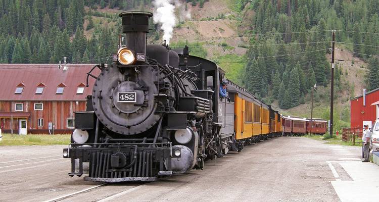 Locomotive à vapeur noire de style ancien émettant de la vapeur à l'arrêt dans une gare rurale avec des voitures jaunes.