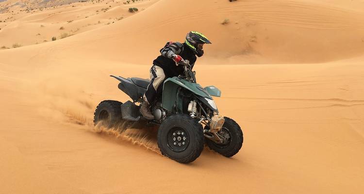 Rider on a quad bike kicks up sand while speeding across orange desert dunes.