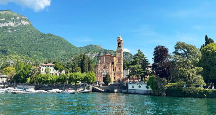 Une église italienne au bord du lac avec un haut clocher située contre des montagnes boisées et un ciel bleu clair, vue depuis l'eau.
