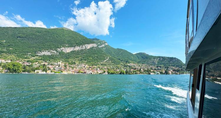 Un lac bleu vibrant avec un village de montagne, photographié depuis le flanc d'un ferry en mouvement sous un ciel lumineux.