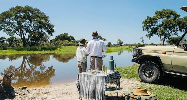 Safari-koppel staat bij een rivieroever picknick-opstelling naast een geparkeerde 4x4 op een heldere blauwe-lucht dag.