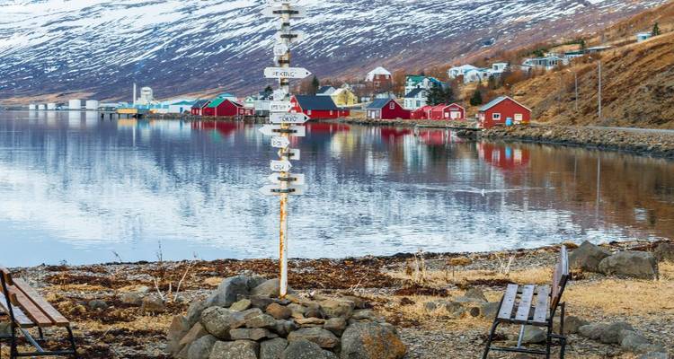 Snow-capped mountains reflected in a calm fjord with colorful red houses lining the shore and a multi-directional signpost in the foreground.