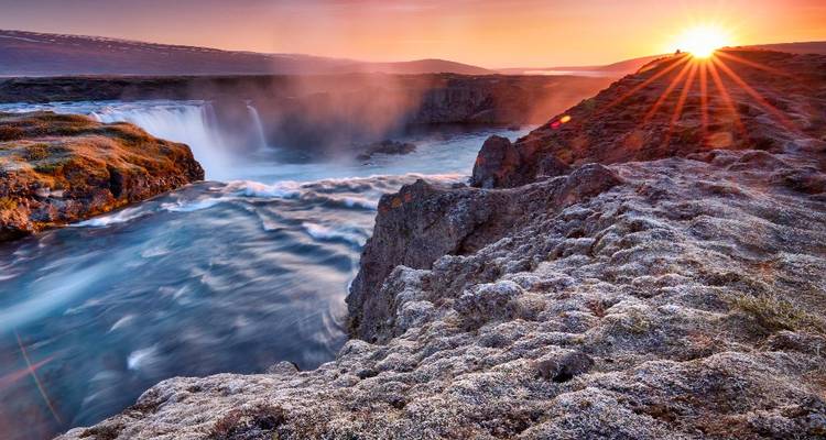 Dramatic waterfall plunging into a misty canyon under a glowing sunrise with starburst light.