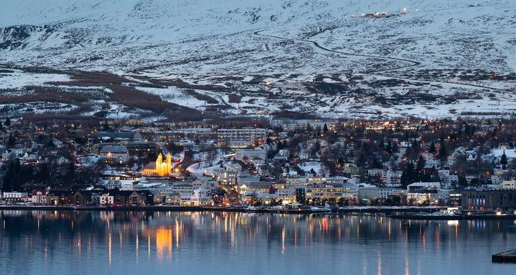 Twilight panorama of a snow-dusted town reflecting lights across a calm fjord.