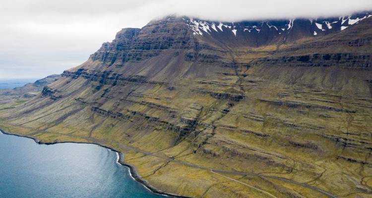 Sheer, layered mountain rising from the sea with cloud-capped summit seen from a drone perspective.
