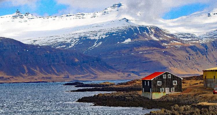 Solitary house with red roof perched on a rocky coastline beneath towering snowy peaks.
