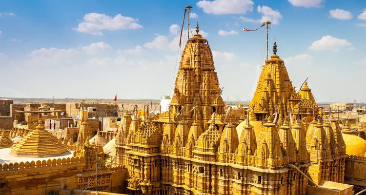 Golden sandstone Jain temple spires rise above the old city with bright blue sky and scattered clouds.